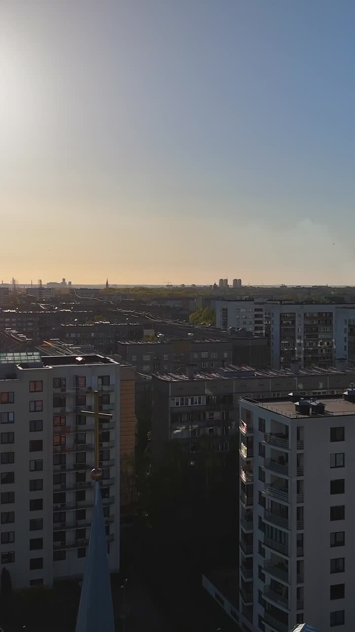 Tall pointed church steeple with a cross rises above Riga apartment buildings in Purvciems Dārzciems district at golden hour, distant cityscape blends into flat Baltic terrain, vertical drone shot