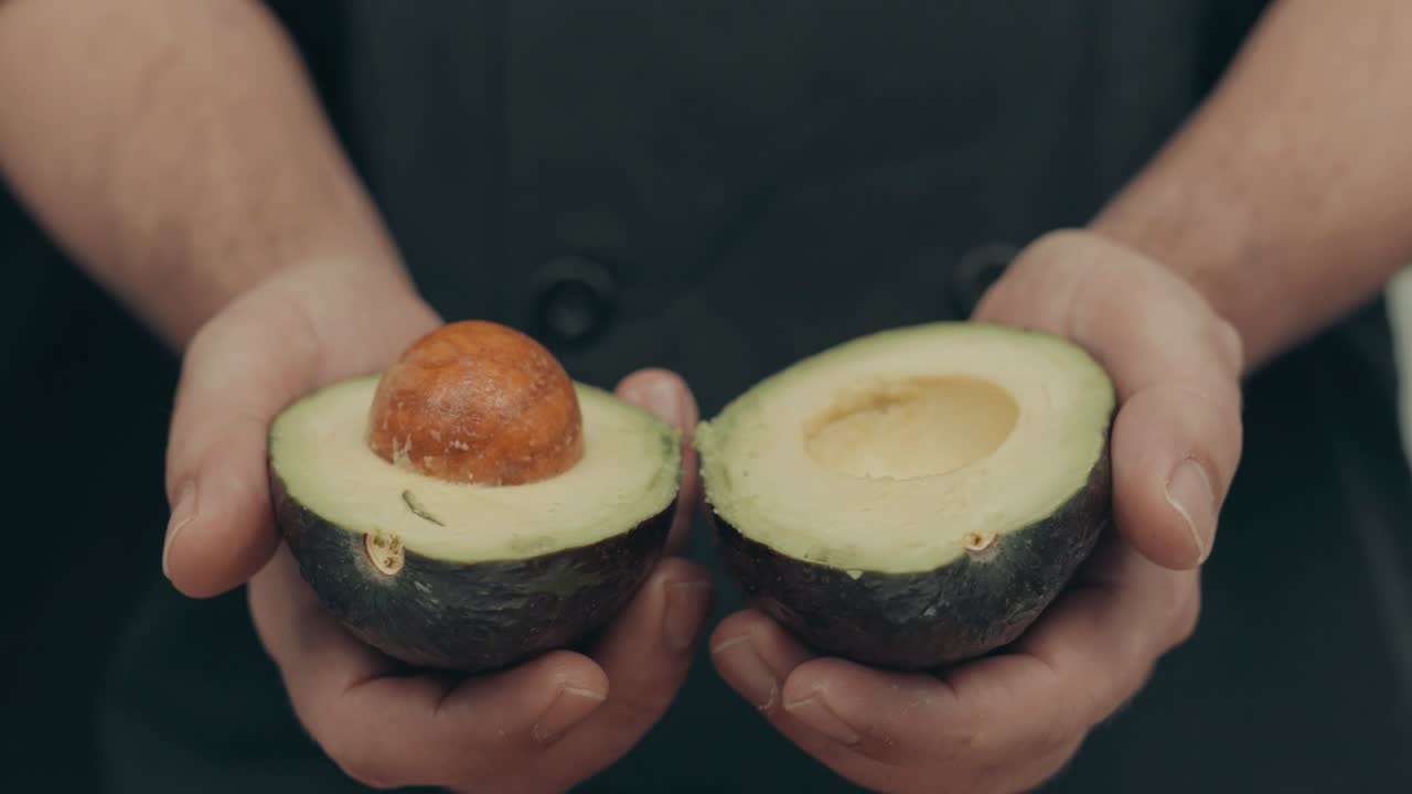 Primo piano di mani maschili che aprono a metà un avocado con il seme marrone all'interno.