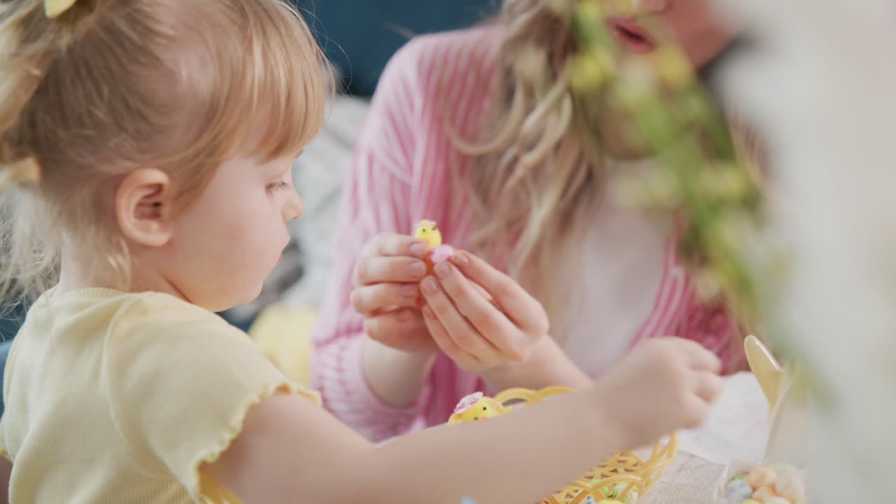 Young girl plays with Easter eggs and bunny toys. Her mother sits nearby, sharing the joyful moment. Soft light highlights their happy expressions. Perfect for family, spring