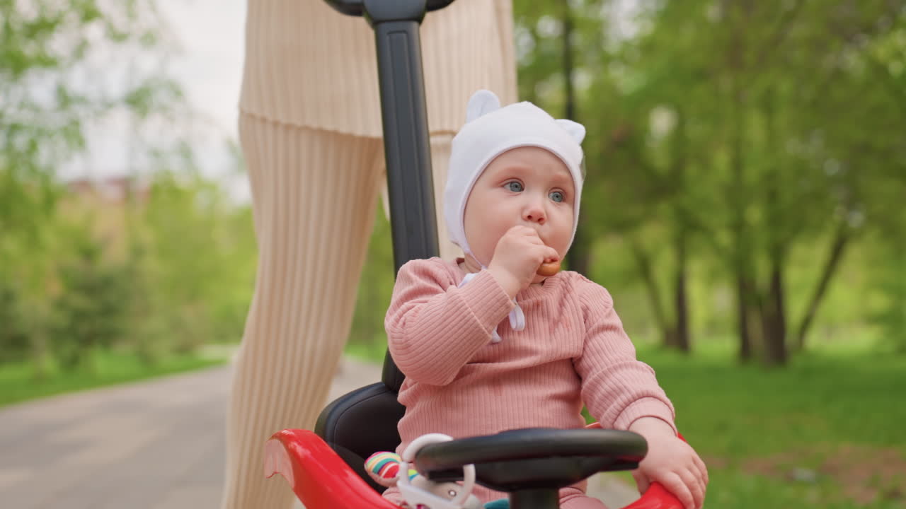 Wide Shot White Mother Pushing Baby Carriage Along Paved Park Path, Seated Baby Chewing Snack In Pushchair, Serene TreeLined Walkway, Minimalist Knit Dress, Relaxed Spring Outing