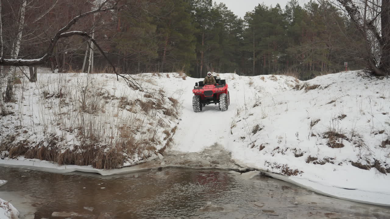 Un quad rosso si lancia ad alta velocità da un pendio innevato in un ruscello con acque basse, creando schizzi e spruzzi, mentre il veicolo fuoristrada si precipita su una sponda ghiacciata in un'esplosione di adrenalina.