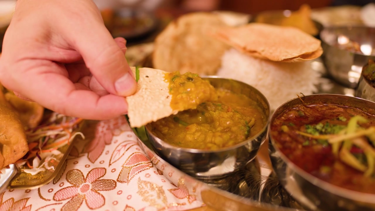 Close-up of a hand dipping crispy papadum into yellow dal stew on a thali.