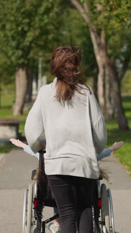 woman pushes daughter with spinal cord injury in wheelchair in city park. loving mom runs on pavement with child enjoying spring nature backside view