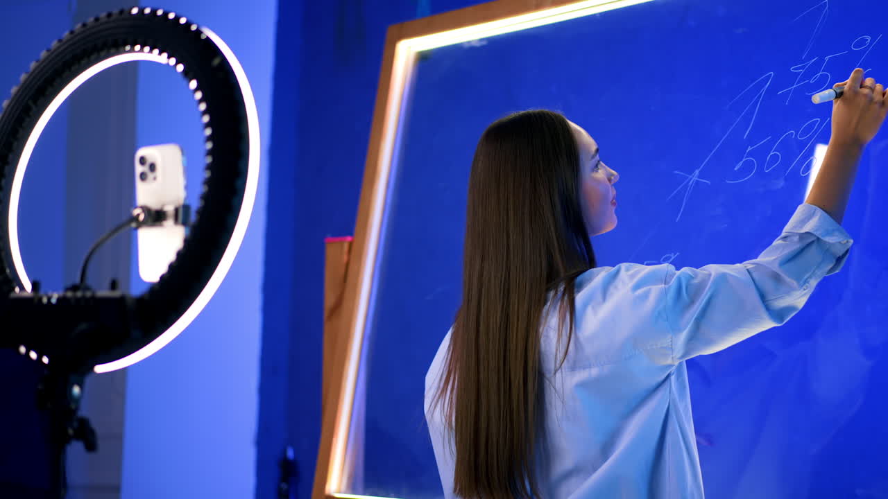 Dark-haired Caucasian lady in a shirt writes on a glass wall with a white marker. Girl turns around to the phone on tripod and talks to camera.