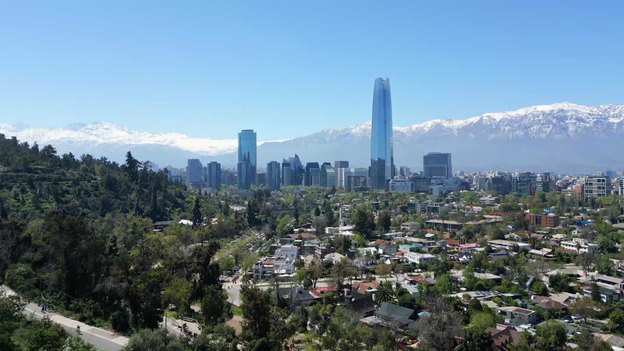 Ripresa aerea con drone che si allontana da Santiago del Cile verso la Costanera Tower e lo skyline del centro con le Ande innevate sotto un cielo sereno diurno