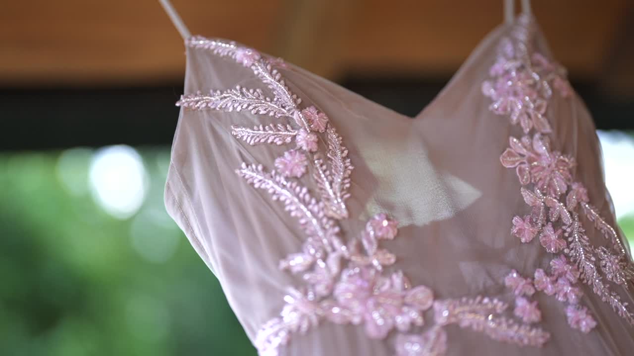 Close-up reveal of bride’s wedding dress hanging, showcasing intricate pastel floral embroidery and sheer, soft fabric before the ceremony begins, prepared and waiting to be worn