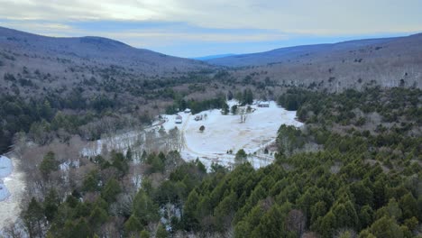 video de drones sobre un bosque de pinos nevados y un prado con casas y montañas distantes en un día nublado de invierno