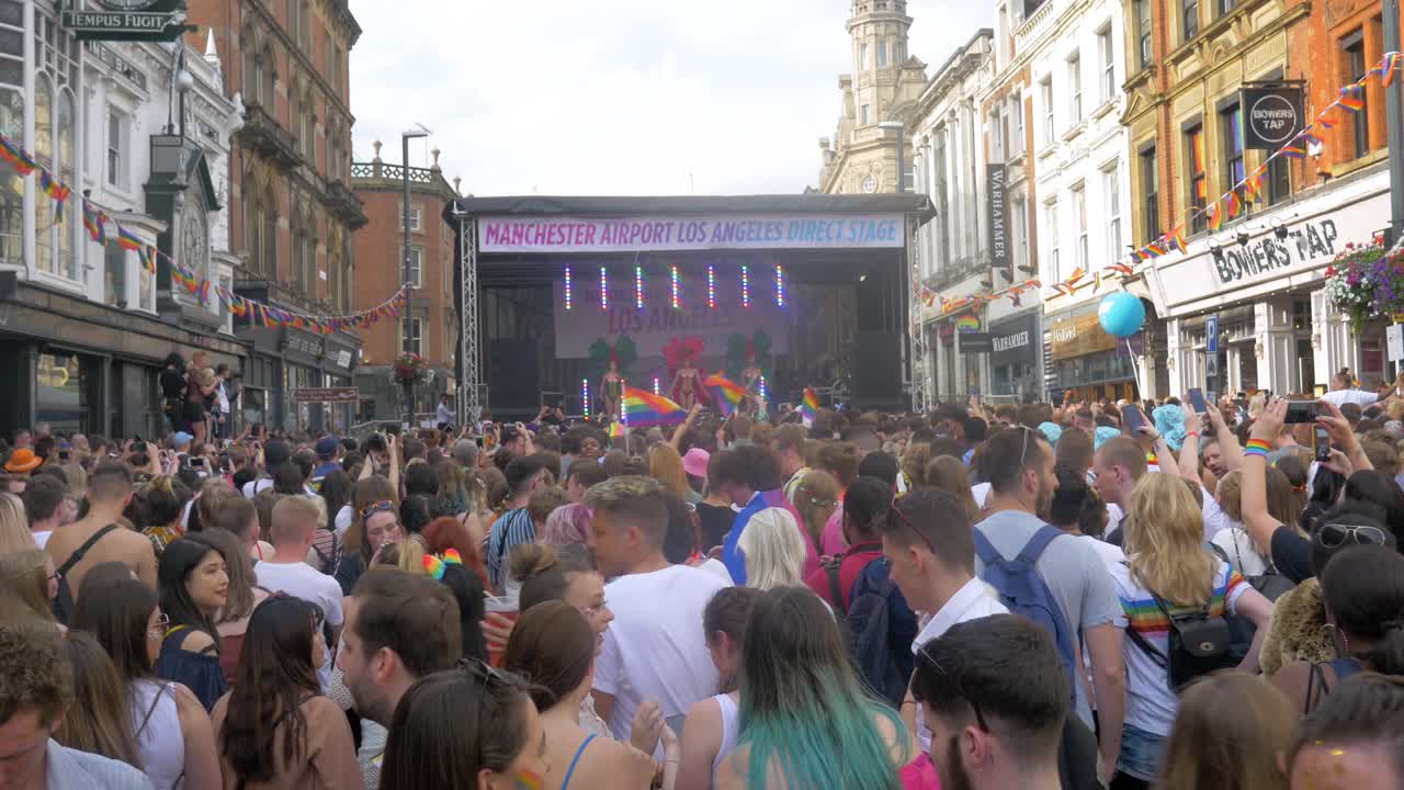 Leeds Pride LGBTQ Festival 2019 shot of the crowd in foreground dancing and waving flags with drag queens on the stage dancing 4K 25p
