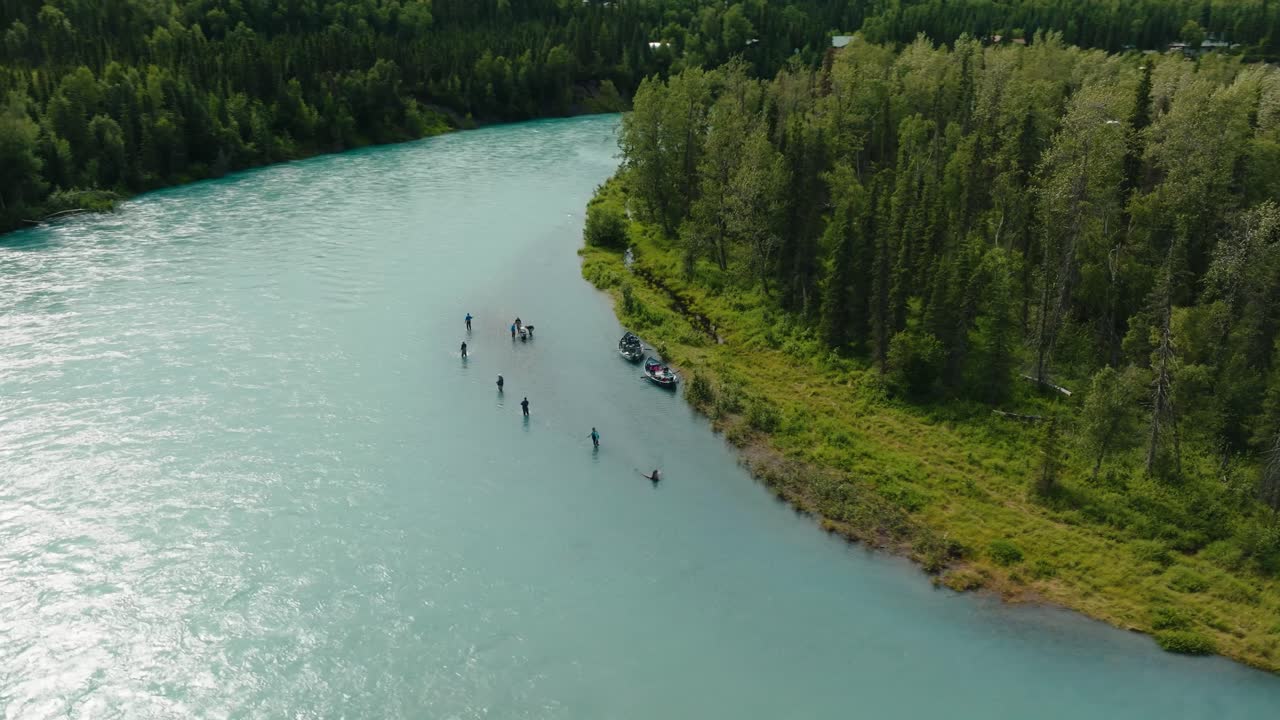 Aerial drone orbit over large group of King Alaskan salmon fisherman, wading and fishing in pristine blue water on lush green riverbed shoreline