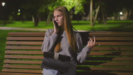 young woman seated on outdoor bench at night, multitasking with phone call and laptop, illuminated by soft park light, red backpack beside her on bench, surrounded by greenery