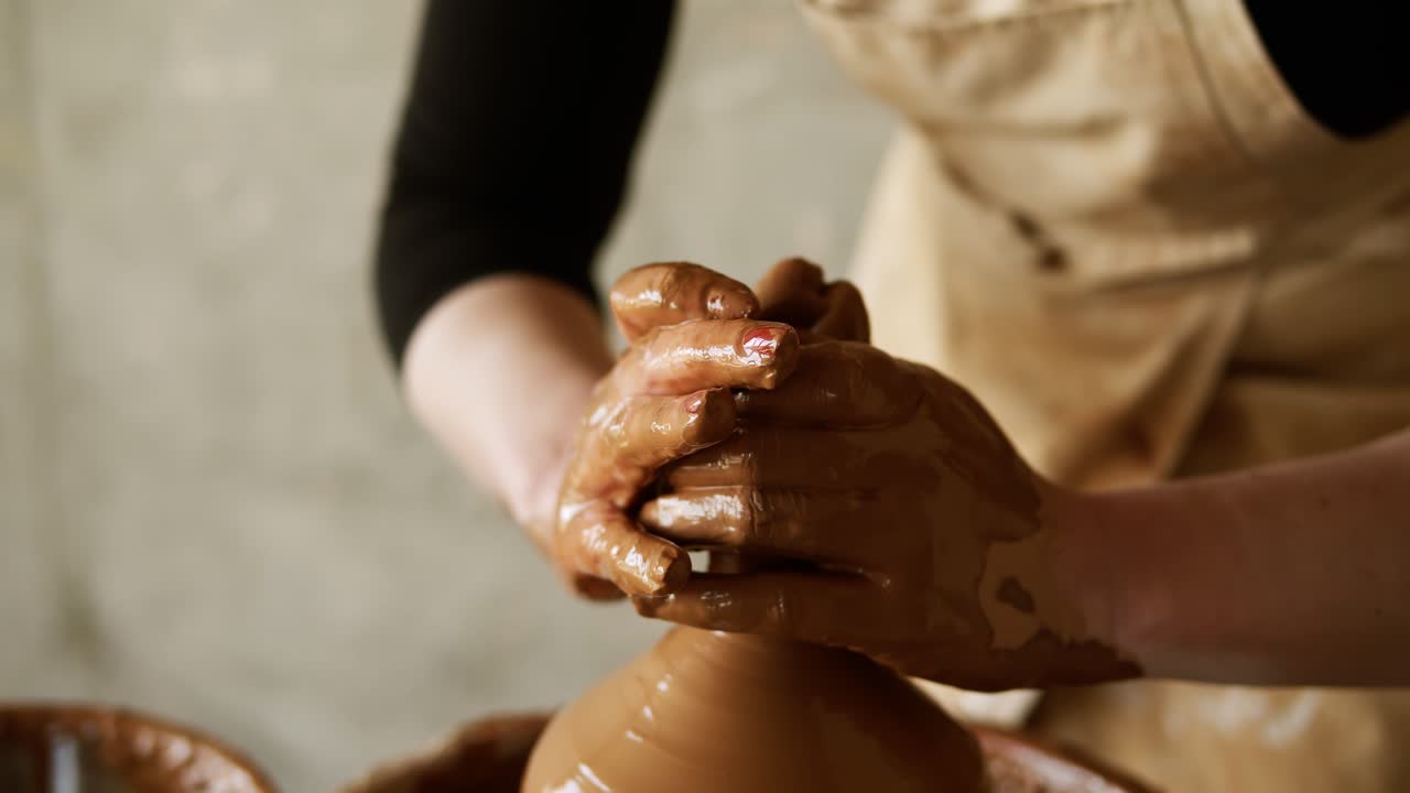 Female potter's hands with red manicure working with wet clay on a pottery wheel making a clay product in a workshop. Unrecognizable female person forming a vase, pulls up the neck