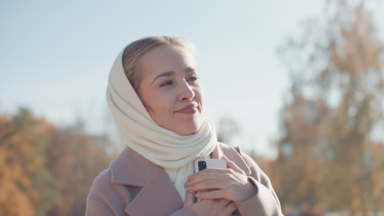 Güneşli bir parkta dışarıda, rahat bir atkı ve paltoya sarınmış, kahvesini tutarken telefonunda mesajları kaydıran, gülümseyen Kafkas kadın. Rahat ve neşeli ifadesiyle, çam ağaçları ve altın sarısı yapraklar arasında doğal bir an.
