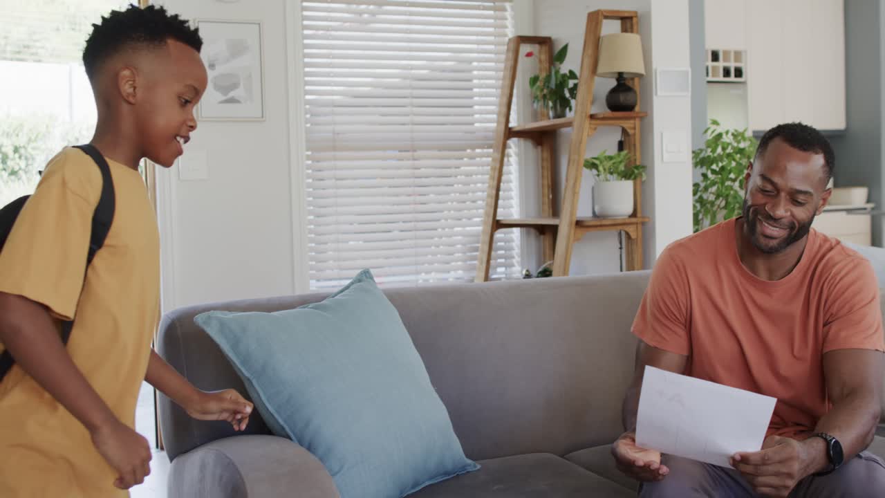 African American dad and son sitting on gray couch, youth entering with backpack, reviewing paper