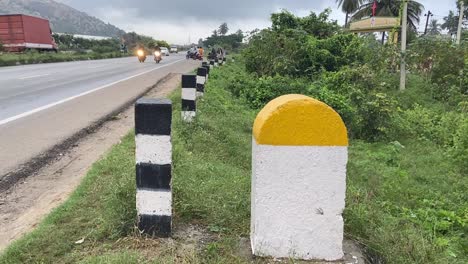 Hito-Amarillo-Y-Blanco-Liso-Con-Carretera-Nacional-Con-Flujo-De-Tráfico-Y-Fondo-De-Plantas-Verdes