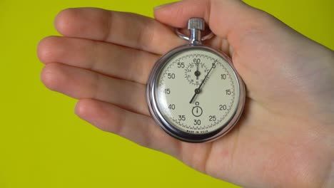 a woman's hand holds an analog stopwatch on a yellow background.