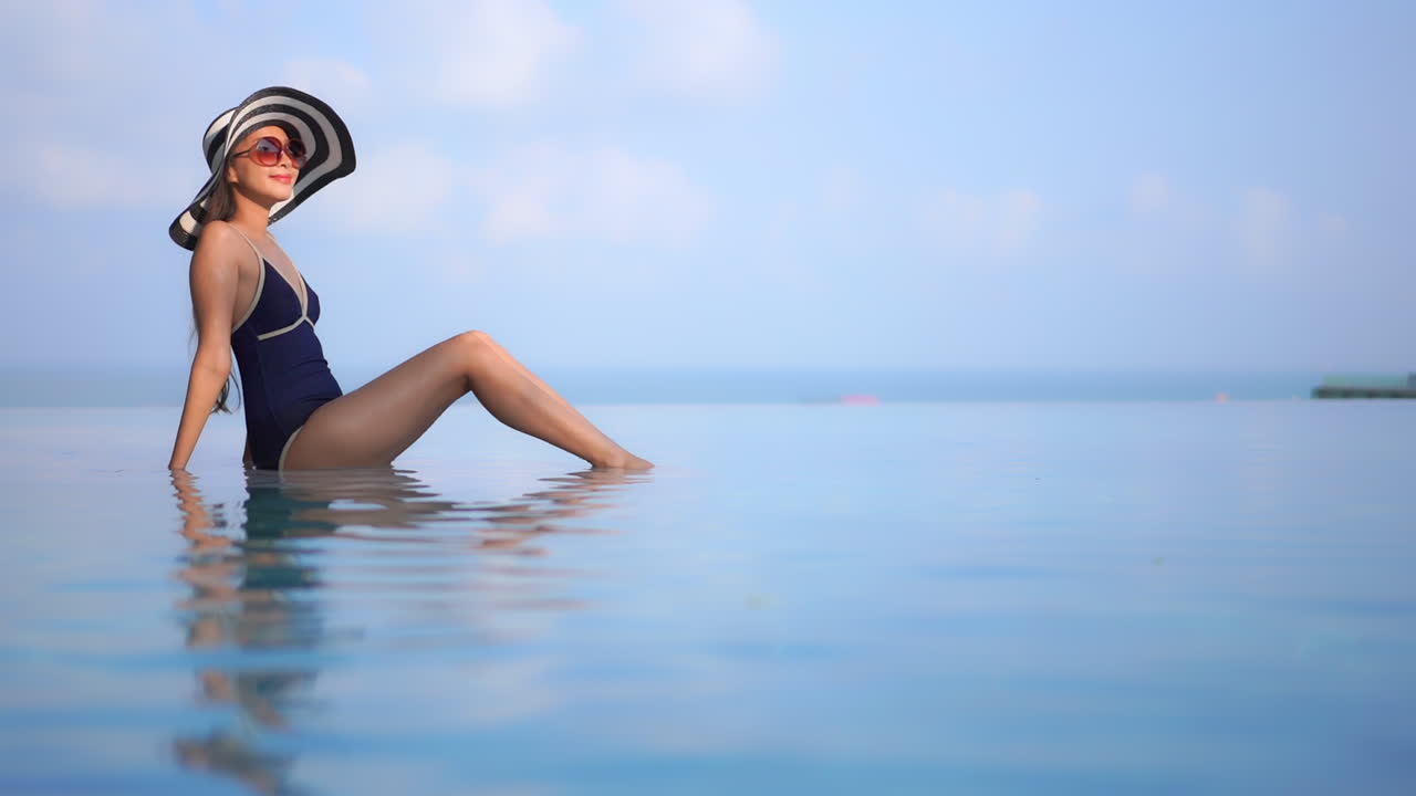 Static wide shot of a beautiful lady sitting on the edge of an infinity pool wearing a beach hat and posing
