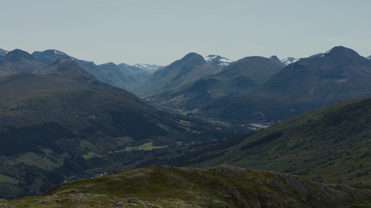 Lang norsk dal skjærer seg mellom mørke fjellsider med fjerne snøflekker under klart middagslys