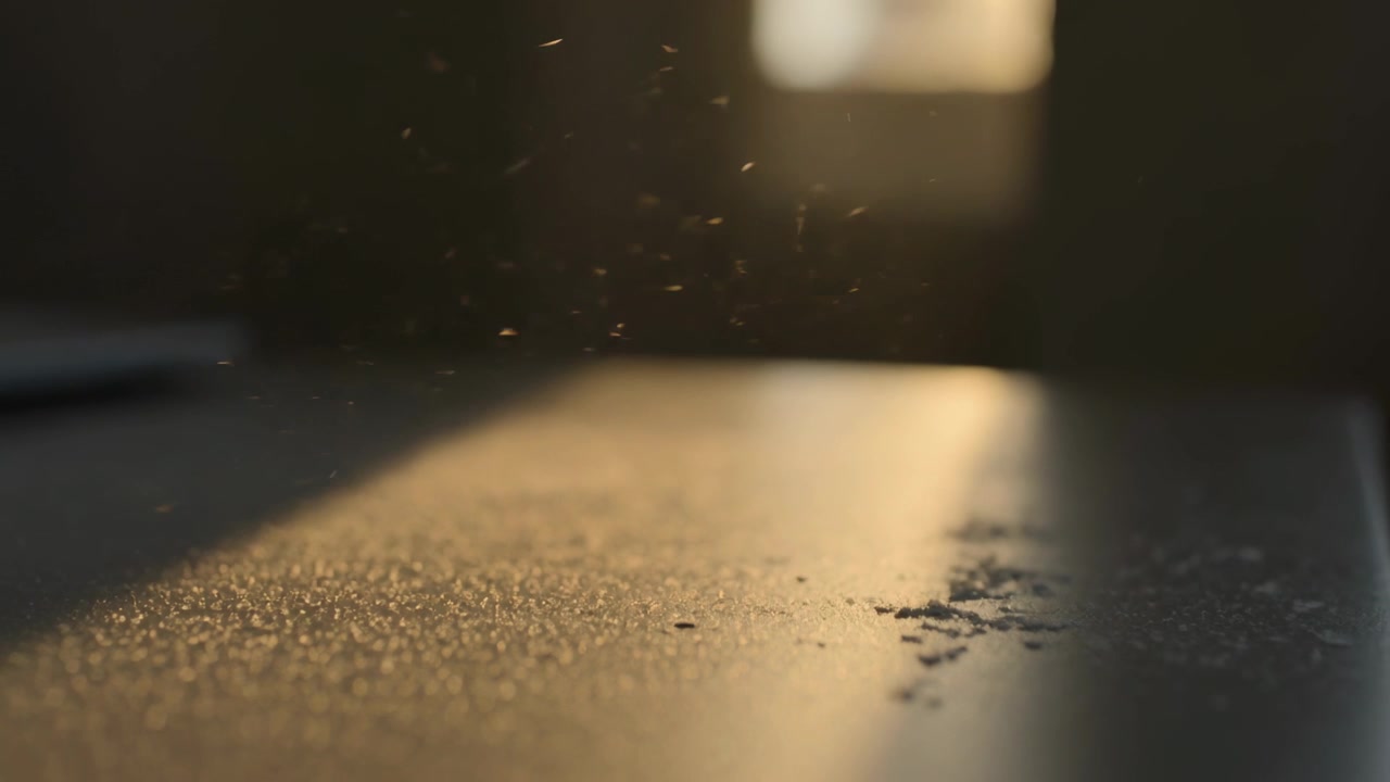 Ein warmer Lichtstrahl durchquert den Raumtisch, Staub schwebt, Krümelhaufen setzen sich nahe dem Fenster-Bokeh ab.