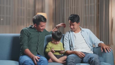 asian gay fathers and their cute son relaxing on couch and using mobile phone and warmly smiling to camera