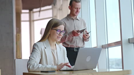 female and male employees in modern office. lady sitting at desk with laptop. male colleague standing behind her with phone and notebook at the panoramic window.