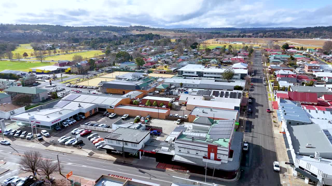 Drone footage glides above a suburban commercial street in Armidale, NSW, Australia, under bright daylight with scattered clouds, revealing rooftops, parked cars, and distant hills