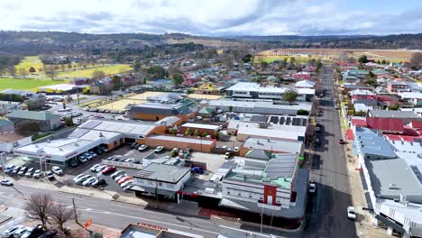 drone footage glides above a suburban commercial street in armidale, nsw, australia, under bright daylight with scattered clouds, revealing rooftops, parked cars, and distant hills