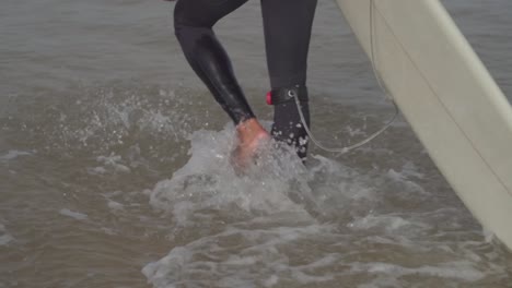 Close-up-shot-of-surfer-walking-into-the-sea-carrying-surfboard,-Slow-motion