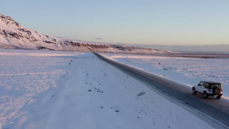 AERIAL:-Flying-besides-Jeep-on-snowy-road-in-Iceland-at-Sunset-with-Sun-Flair-Winter,-Sun,-Arctic