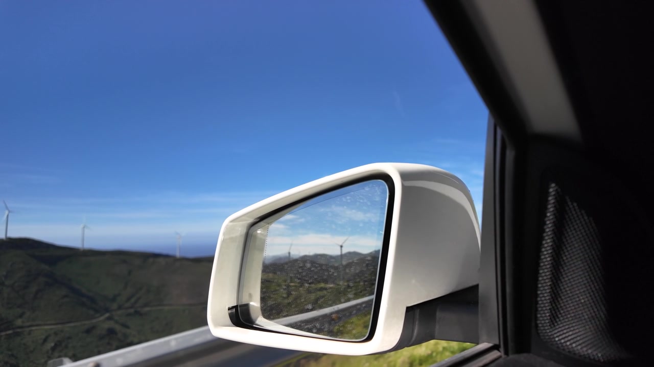 Specchietto retrovisore dell'auto che riflette turbine eoliche montane sopra il mare