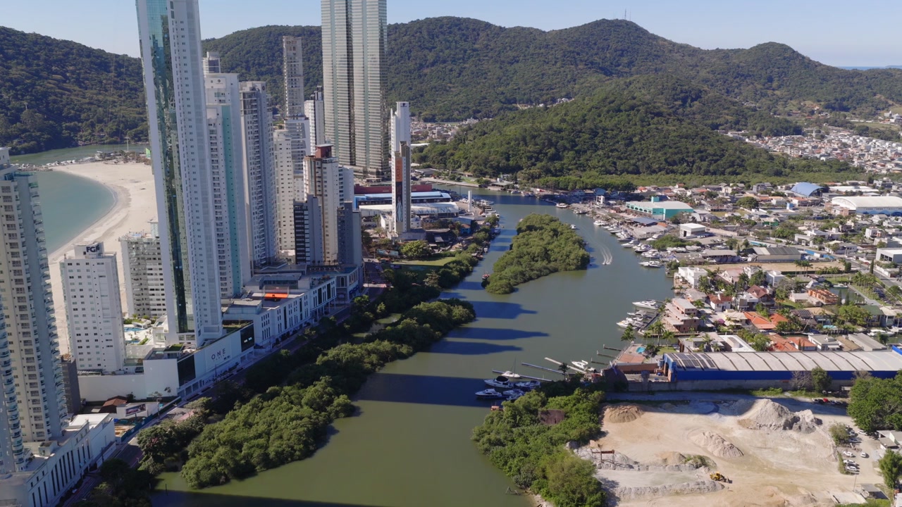Aerial view of high-rise buildings beside Rio Palmital river with green hills in background, Balneário Barra do Sul, Santa Catarina, Brazil.