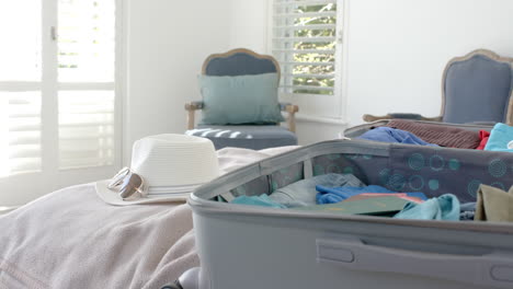 Close-up-of-suitcase,-hat-and-sunglasses-on-bed-in-bedroom,-slow-motion