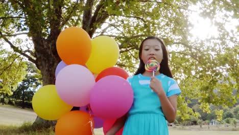Portrait-of-smiling-girl-holding-balloons-and-lollypop-in-the-park