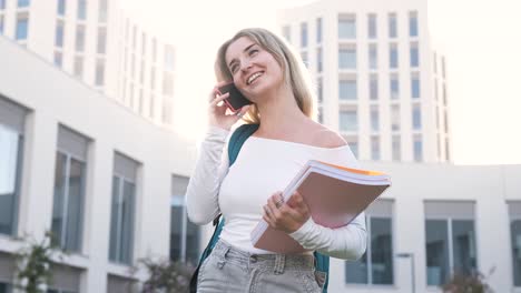 Smiling woman with books talking on cellphone outside college