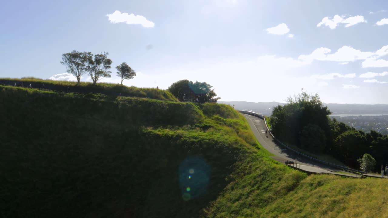una toma de mano de la cumbre del volcán mount eden en auckland, nueva zelanda, con árboles en la parte superior y un camino en el lado, con turistas ascendiendo al volcán en una tarde clara y azul
