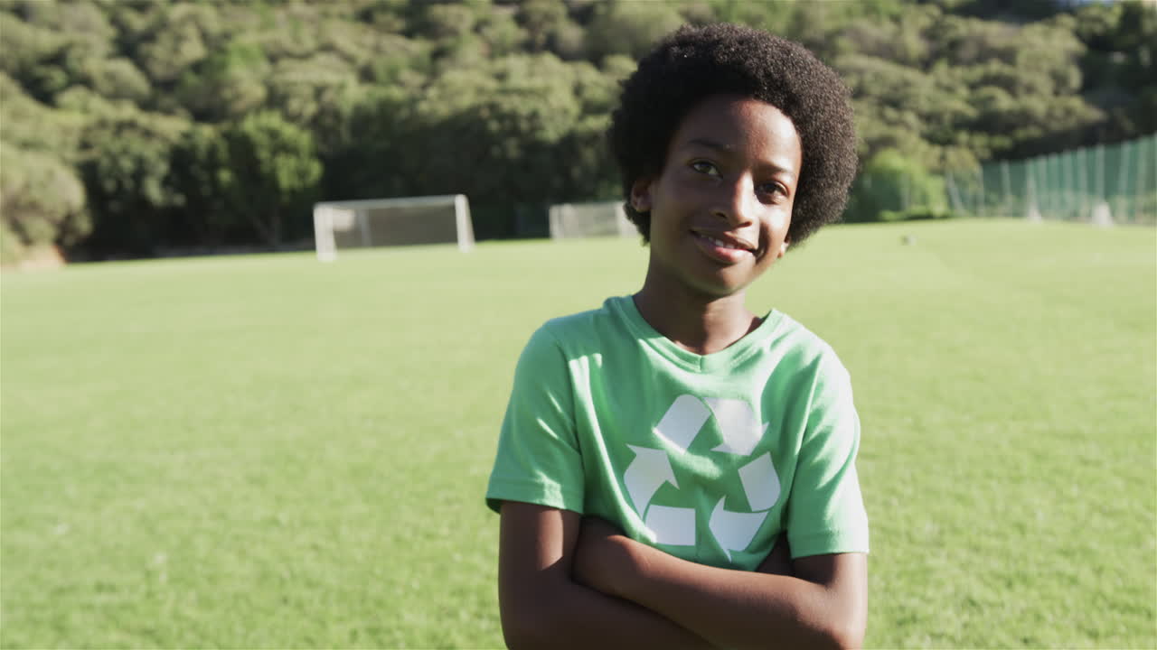 niño biracial se para con confianza en un campo de fútbol con una camiseta de reciclaje verde, con espacio de copia