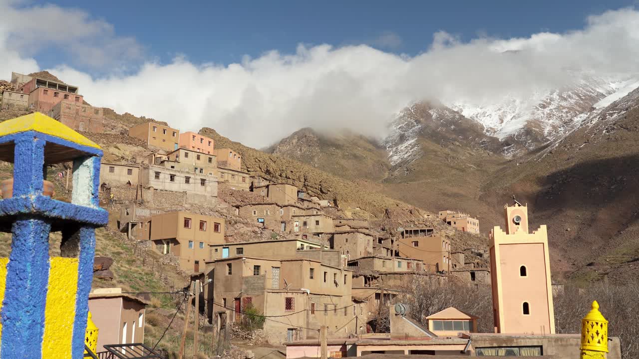 In the small courtyard of the mountain village, stone houses are grouped closely, with the towering Toubkal peaks providing an imposing yet serene view from the heart of the village in Morocco