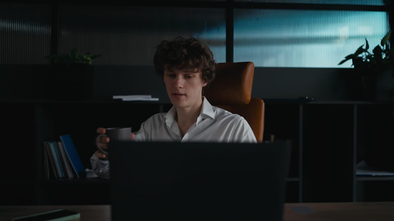 A young guy with curly hair in a white shirt sits at a table and works on a laptop while drinking coffee in the office