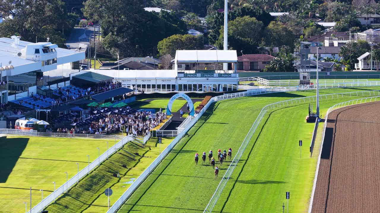 Drone footage captures jockeys riding horses along a green turf track at an outdoor racecourse, with crowds watching under clear daylight