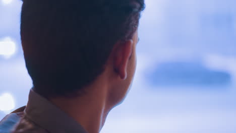 young man in profile sitting near window, looking sideways with thoughtful expression, waiting or reflecting, natural light illuminating face and hair, background blurred, tones, shirt visible