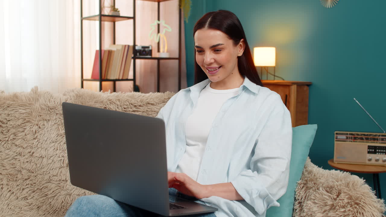 Young woman freelancer sits on sofa working with laptop focused and typing concentrated on project
