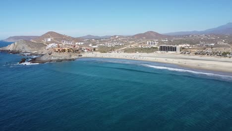 aerial shot over the beautiful resort of cerritos beach in mexico with a view of the turquoise sea with calm waves, hotel buildings, many beachgoers and dry landscape on a cloudless day