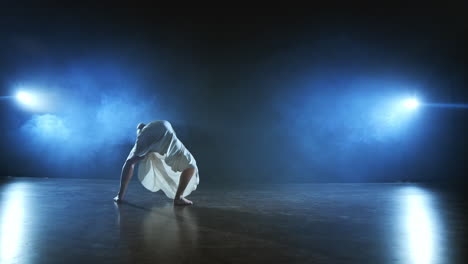 a young girl in a white dress dances contemporary in slow motion on the stage with smoke in spotlights. camera stands behind her filming in a full shot.