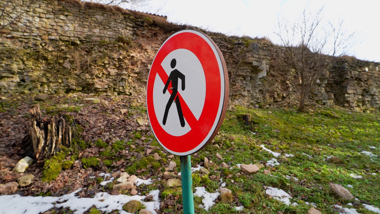 Warning sign forbidding pedestrian access near a crumbling historic stone wall, with loose rocks and winter ground highlighting danger, instability, and restricted passage.