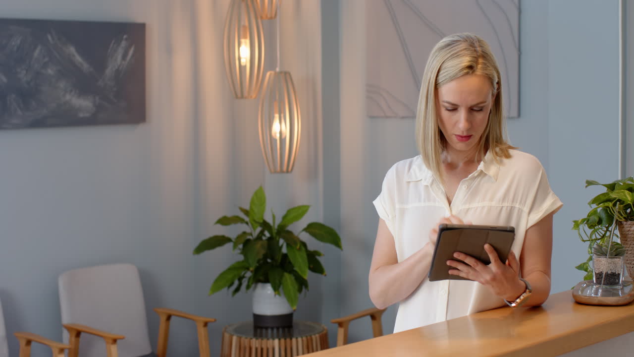 A middle-aged blonde Caucasian woman with fair skin stands at reception using tablet