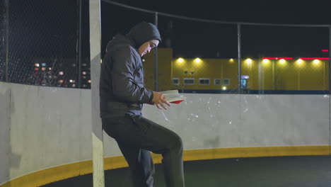 athlete in hoodie leaning against metallic goalpost outdoors at night flipping pages of book with soccer ball gently rolling underfoot, illuminated by warm urban city lights