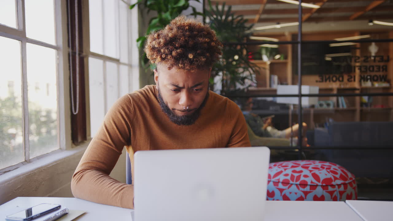 blogger masculino negro del milenio usando una computadora portátil un teléfono inteligente en un escritorio en una oficina creativa, de cerca