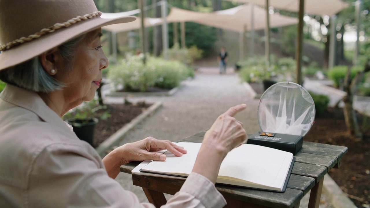 Donna anziana con un cappello che tiene un taccuino, apre le pagine a un tavolo da giardino, tocca una cupola, con spazio per il testo