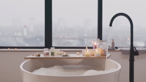 camera focuses on a white bathtub filling with water and foam and wooden table with bathroom elements while a tap is pouring water