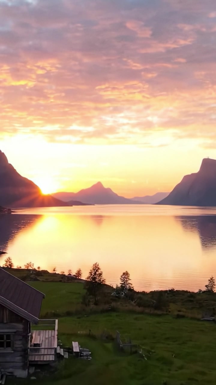 En fredfyldt solnedgang over vand med majestætiske bjerge og en rustik hytte i forgrunden, der indfanger skønheden i naturens ro ved skumringstid.