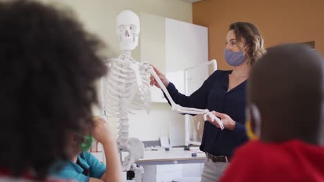 maestra que usa una máscara facial usando un modelo de esqueleto humano para enseñar a los estudiantes en clase